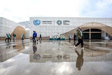 Mehrere Menschen passieren das COP29-Gebäude mit UN-Klimawandel-Logo, nasser Boden reflektiert ihre Bewegungen.