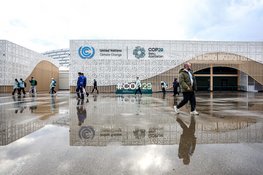 Mehrere Menschen passieren das COP29-Gebäude mit UN-Klimawandel-Logo, nasser Boden reflektiert ihre Bewegungen.
