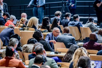 John Podesta, Sonderbeauftragter des US-Präsidenten für das Klima, im Gespräch mit Ali Mohamed, Klimabotschafter der kenianischen Regierung. Das Forum der COP stellt durch die Teilnahme aller Staaten sowie die Präsenz der Zivilgesellschaft und der beobachtenden Wissenschaft eine Verhandlungsarena von großem Wert dar. Mehrere Delegierte in einer Konferenzhalle, vertieft in Gespräche, während sie an einer internationalen Veranstaltung teilnehmen. Im Hintergrund sind Kameras und weitere Teilnehmer zu sehen.