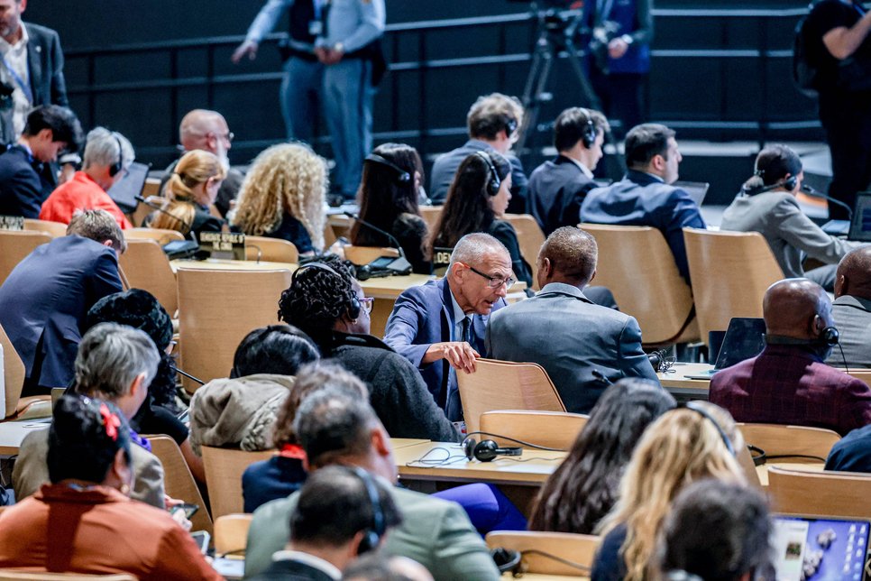 Mehrere Delegierte in einer Konferenzhalle, vertieft in Gespräche, während sie an einer internationalen Veranstaltung teilnehmen. Im Hintergrund sind Kameras und weitere Teilnehmer zu sehen.