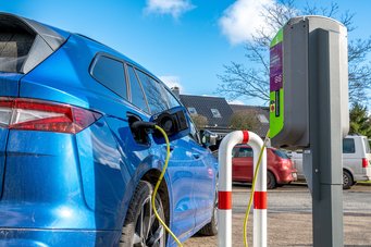 Ein blaues Elektroauto ist an eine Ladestation angeschlossen. Im Hintergrund sind Häuser und geparkte Autos zu sehen, während der Himmel klar ist.