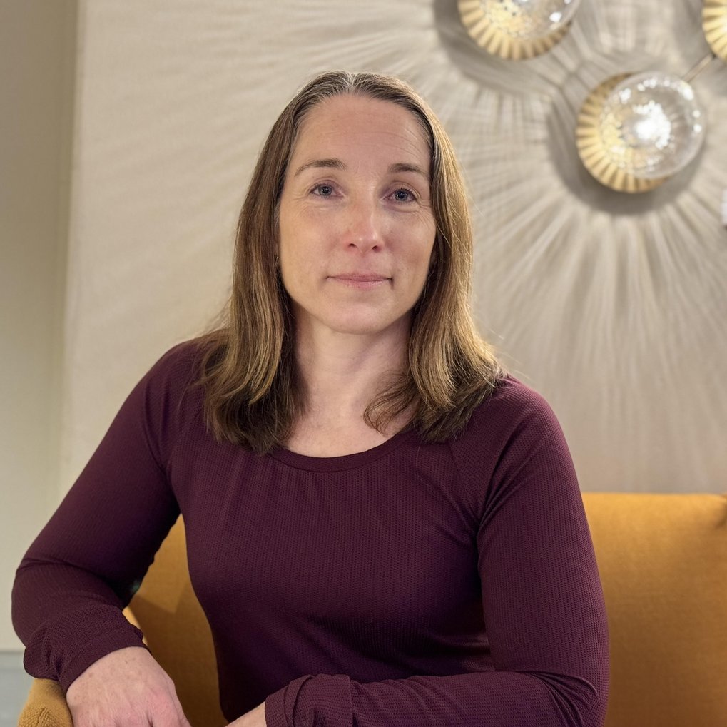 Eine Frau in einem bordeauxfarbenen Pullover sitzt entspannt auf einem gelben Sofa, im Hintergrund sind dekorative Wandlampen zu sehen.