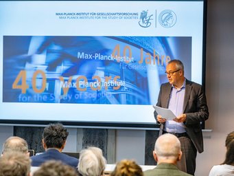 Eine Veranstaltung zum 40-jährigen Jubiläum des Max-Planck-Instituts für Gesellschaftsforschung, bei der ein Redner vor einer Präsentation spricht. Jens Beckert hält eine Präsentation im Max-Planck-Institut für Gesellschaftsforschung, das sein 40-jähriges Bestehen feiert. Die Folie zeigt das Jubiläumslogo und die Institutsnamen in Deutsch und Englisch. Publikum im Vordergrund.