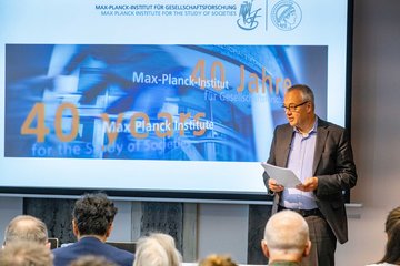 Eine Veranstaltung zum 40-jährigen Jubiläum des Max-Planck-Instituts für Gesellschaftsforschung, bei der ein Redner vor einer Präsentation spricht. Jens Beckert hält eine Präsentation im Max-Planck-Institut für Gesellschaftsforschung, das sein 40-jähriges Bestehen feiert. Die Folie zeigt das Jubiläumslogo und die Institutsnamen in Deutsch und Englisch. Publikum im Vordergrund.