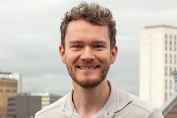 Eine Person trägt ein hellgraues Hemd und steht auf einer Dachterrasse, umgeben von modernen Gebäuden, während der Himmel bewölkt ist. Im Hintergrund sind mehrstöckige Gebäude zu sehen.