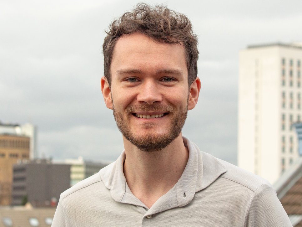 Eine Person trägt ein hellgraues Hemd und steht auf einer Dachterrasse, umgeben von modernen Gebäuden, während der Himmel bewölkt ist. Im Hintergrund sind mehrstöckige Gebäude zu sehen.