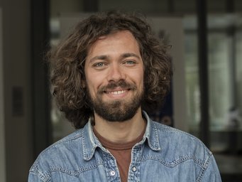 Ein Mann mit lockigem Haar, das seine Schultern bedeckt, trägt ein Jeanshemd über einem braunen T-Shirt. Hintergrund unscharf. Das Foto zeigt Matteo Marenco, der zur Europäischen Kommission gewechselt ist.