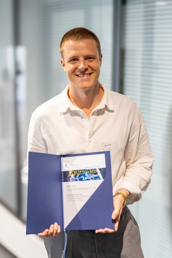 Eine Person in einem hellen Hemd zeigt eine Auszeichnung in einer blauen Mappe. Auf der Urkunde steht „Journal Article Prize 2023“ und „Dustin Voss“. Der Hintergrund ist ein modernes Büro mit Glaswänden.