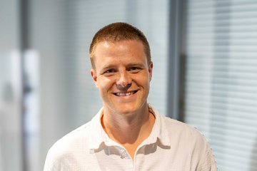 Eine Person in einem hellen Hemd zeigt eine Auszeichnung in einer blauen Mappe. Auf der Urkunde steht „Journal Article Prize 2023“ und „Dustin Voss“. Der Hintergrund ist ein modernes Büro mit Glaswänden.