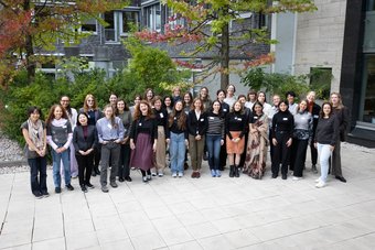 Eine Gruppe von Personen hat sich im Freien versammelt, mit Grünflächen und Bäumen im Hintergrund, und steht auf einem gepflasterten Bereich in der Nähe einer modernen Gebäudefassade.