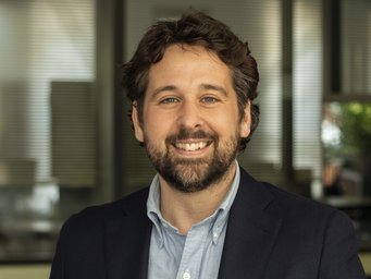 Eine Person trägt ein dunkelblaues Sakko über einem hellblauen Hemd und steht vor einer Reihe von Bürofenstern. Im Hintergrund sind verschwommene Bürogegenstände zu erkennen.