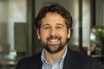 Eine Person trägt ein dunkelblaues Sakko über einem hellblauen Hemd und steht vor einer Reihe von Bürofenstern. Im Hintergrund sind verschwommene Bürogegenstände zu erkennen.
