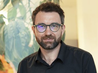 Mann in schwarzem Hemd, steht vor einer großen grünen Pflanze in einem hellen Raum.