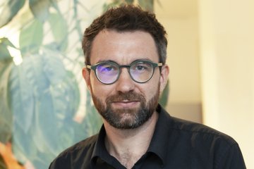 Mann in schwarzem Hemd, steht vor einer großen grünen Pflanze in einem hellen Raum.