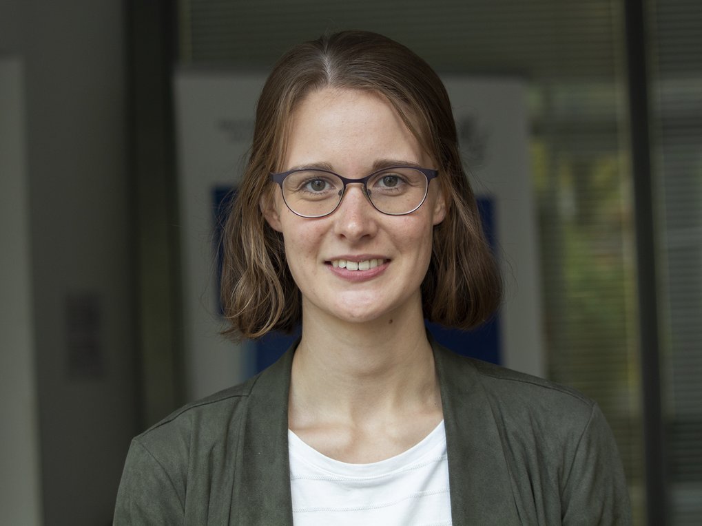 Eine Frau mit schulterlangem, braunem Haar trägt eine graue Jacke und ein weißes T-Shirt. Sie steht vor einem modernen Bürogebäude mit Glasfronten.