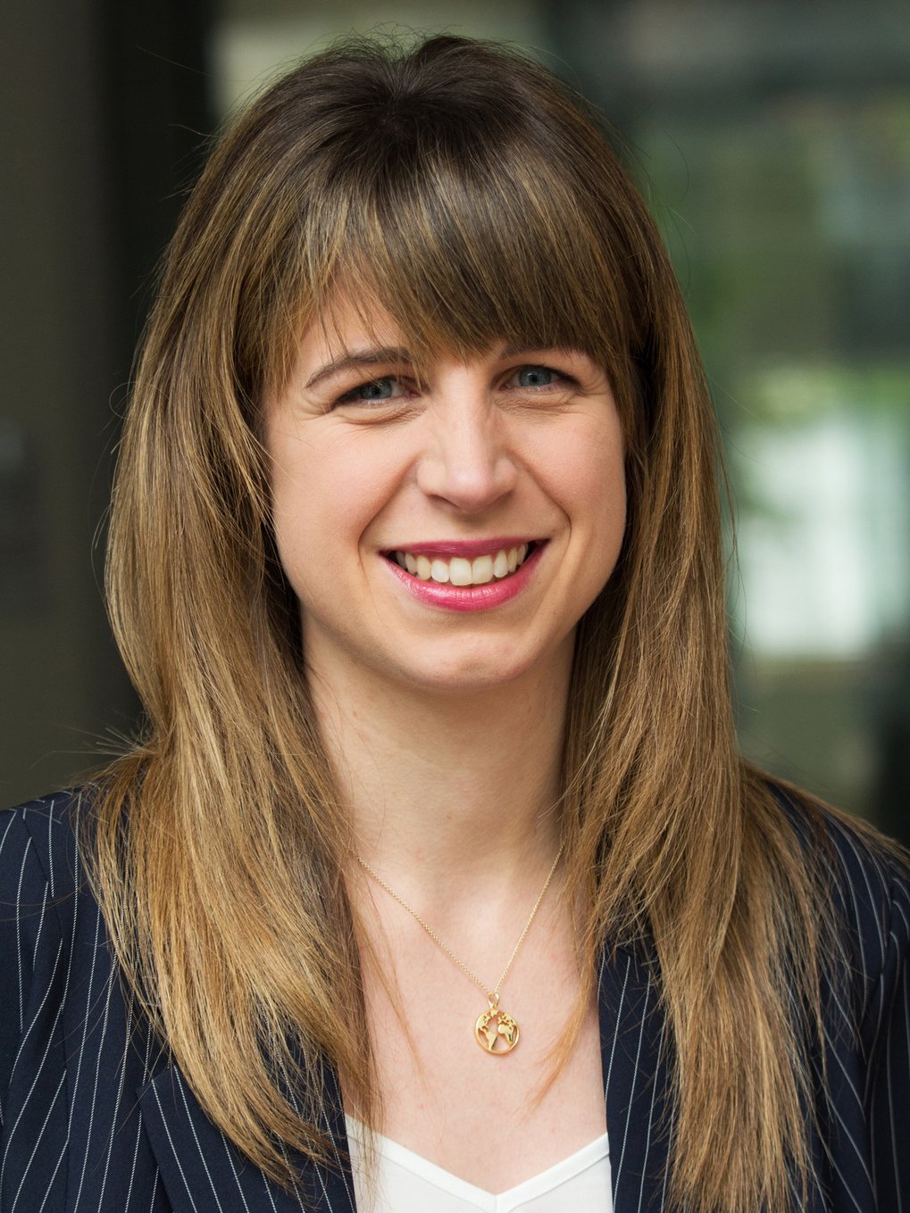 Eine Person mit langen, braunen Haaren, die einen eleganten, dunkelblauen Blazer mit Nadelstreifen und eine goldene Halskette mit einem runden Anhänger trägt.