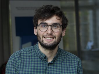 Person trägt ein grün-blau kariertes Hemd. Der Hintergrund zeigt ein Fenster mit strukturiertem Glas und unscharfe Details.