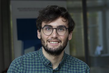 Person trägt ein grün-blau kariertes Hemd. Der Hintergrund zeigt ein Fenster mit strukturiertem Glas und unscharfe Details.