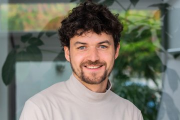 Eine Person mit lockigem Haar trägt einen weißen Rollkragenpullover, im Hintergrund sind grüne Pflanzenblätter sichtbar.