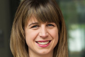 A person with long brown hair wearing an elegant dark blue pinstriped blazer and a gold necklace with a round pendant.