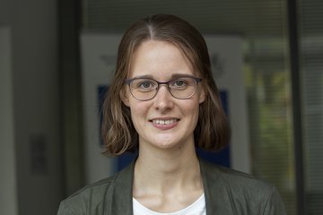 A woman with shoulder-length brown hair is wearing a gray jacket and a white T-shirt. She is standing in front of a modern office building with glass facades.