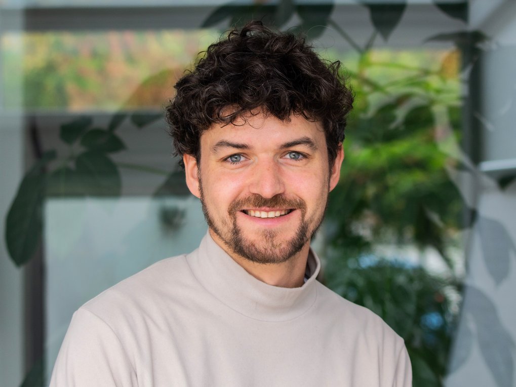 A person with curly hair is wearing a white turtleneck sweater, with green plant leaves visible in the background.