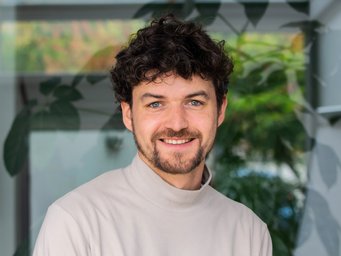 A person with curly hair is wearing a white turtleneck sweater, with green plant leaves visible in the background.