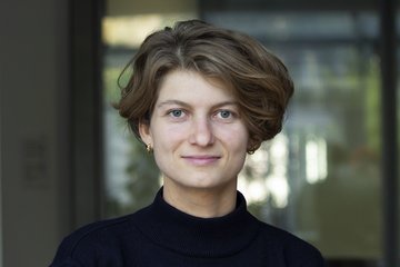 Frau mit hellbrauner Kurzhaarfrisur, in einem dunklen Pullover und goldenen Ohrringen. Hintergrund leicht verschwommen mit Glasstruktur.