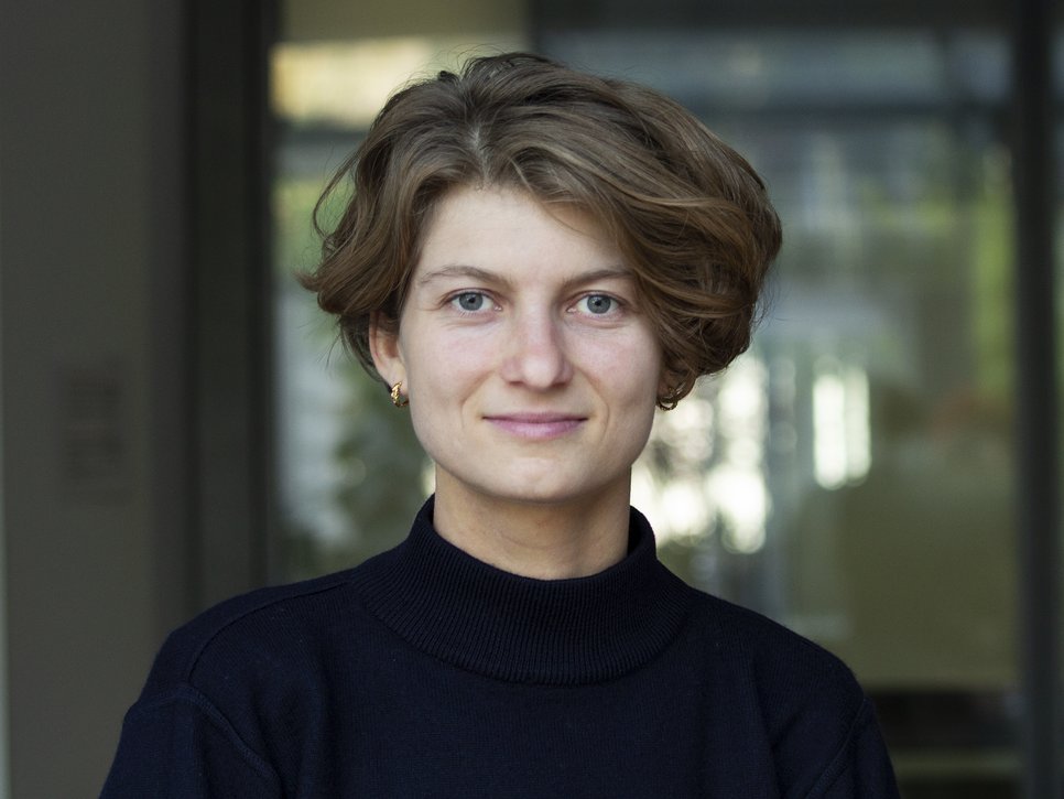 Frau mit hellbrauner Kurzhaarfrisur, in einem dunklen Pullover und goldenen Ohrringen. Hintergrund leicht verschwommen mit Glasstruktur.