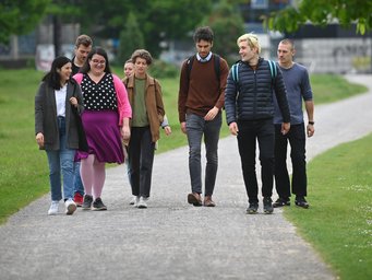 Eine Gruppe aus sieben Personen spaziert einen breiten Kiesweg entlang, flankiert von grünen Wiesen und vereinzelten Bäumen. Sie tragen legere Kleidung und einige haben Rucksäcke dabei.