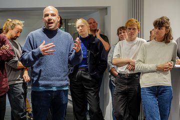 Mehrere Menschen in Freizeitkleidung stehen in einem modernen Raum, während ein Sprecher gestikuliert und Aufmerksamkeit erhält.