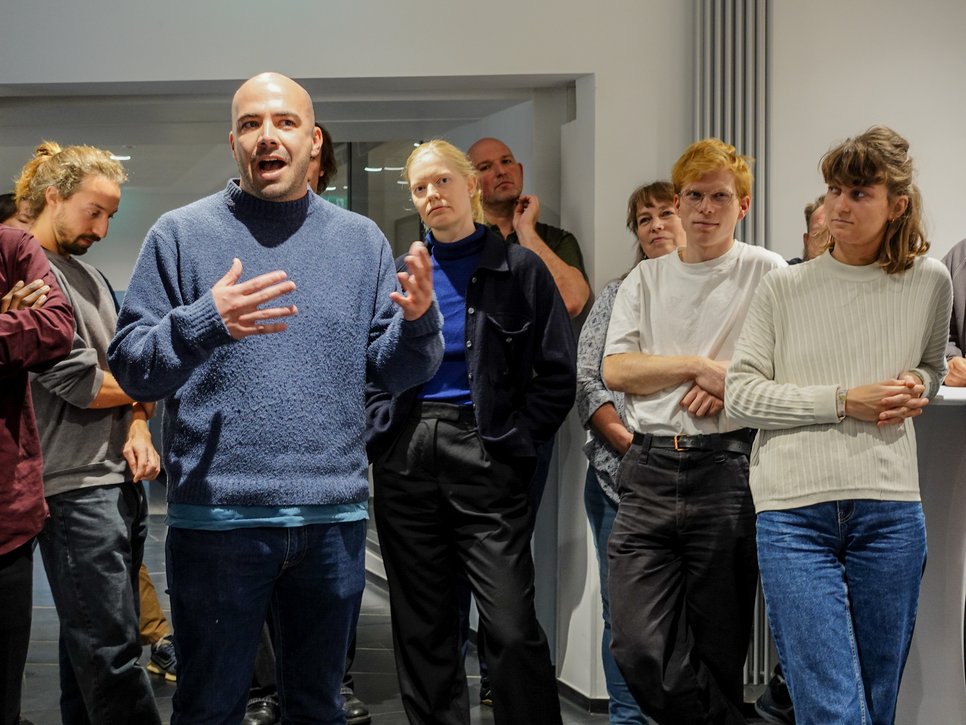 Several people in casual clothing are standing in a modern room while a speaker gestures and gets attention.