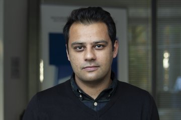 Person is wearing a dark blue sweater over a plaid shirt. A window and a glass door can be seen in the background.