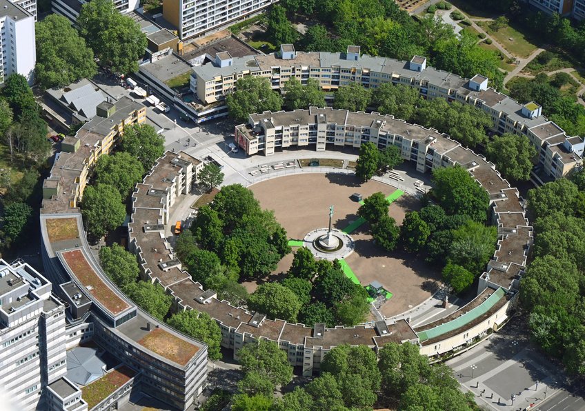 Die Geschichte des Wohnungskomplexes am Berliner Mehringplatz zeigt, was passieren kann, wenn Investoren Wohnraum vor allem nutzen, um Rendite zu erzielen. Luftaufnahme eines kreisförmigen Wohnkomplexes mit einem zentralen Platz und einer Statue, umgeben von Bäumen und Straßen.