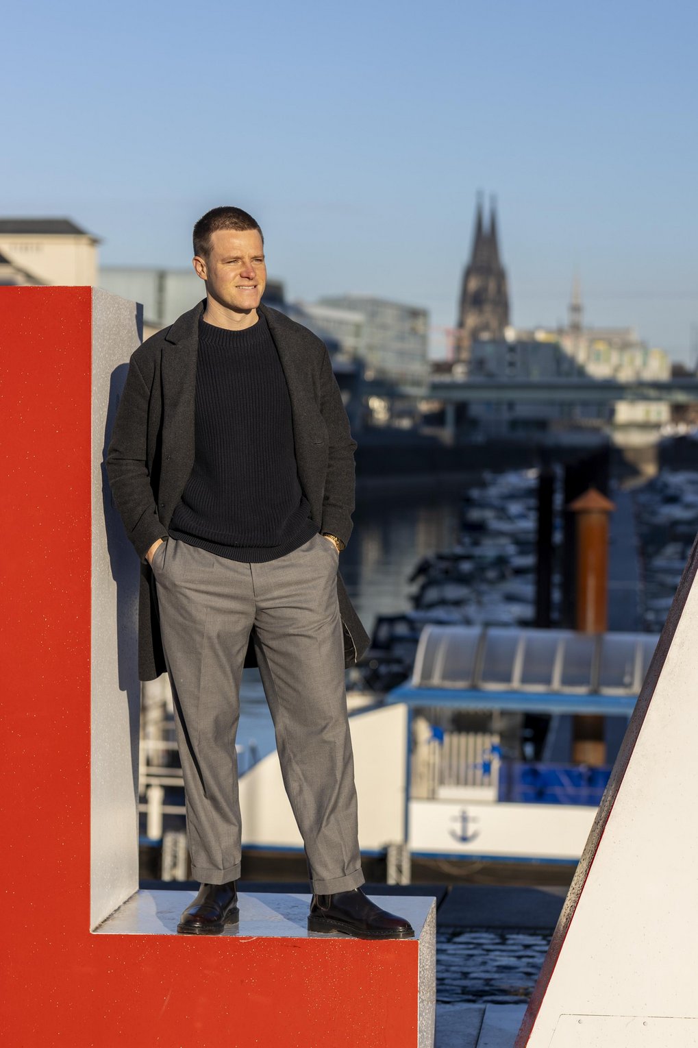 Im Vordergrund steht eine Person an einem roten Objekt, während im Hintergrund Boote im Hafen und der Kölner Dom zu sehen sind.