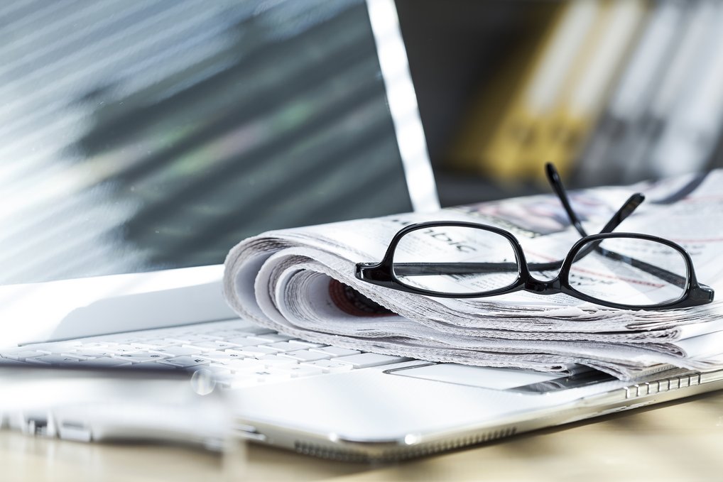 Eine Brille liegt auf einer Zeitung, die auf der Tastatur eines aufgeklappten Laptops liegt.
