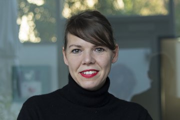 Das Foto zeigt das Porträt eines Individuums der Gattung Homo sapiens, Unterart Homo sapiens intellectualis. Weitere Merkmale wie Lippenstift, Schminke und Frisur deuten auf weibliche Genderidentifikation hin. Das Individuum trägt einen schwarzen Roll-Pullover und steht oder sitzt lächelnd vor einem vermutlich gläsernen spiegelnden Hintergrund in die Kamera.