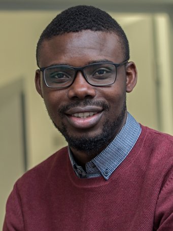 Ein lächelnder Mann mit Brille und burgunderrotem Pullover, darunter ein Hemd mit Kragen, sitzt in einem Innenraum.