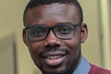 Ein lächelnder Mann mit Brille und burgunderrotem Pullover, darunter ein Hemd mit Kragen, sitzt in einem Innenraum.