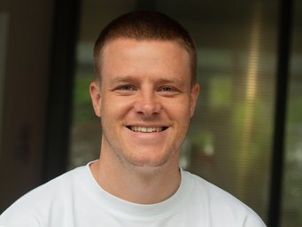 Lächelnde Person in einem weißen Hemd vor einem unscharfen Hintergrund. Der Ton ist warm und zugänglich und vermittelt Freundlichkeit und Positivität.