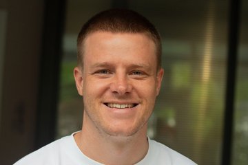 Lächelnde Person in einem weißen Hemd vor einem unscharfen Hintergrund. Der Ton ist warm und zugänglich und vermittelt Freundlichkeit und Positivität.