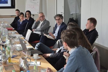 Eine Gruppe von Fachleuten sitzt um einen Konferenztisch herum und diskutiert. Auf dem Tisch stehen Laptops, Notizbücher und Getränke, was auf eine gemeinsame Besprechung hindeutet.
