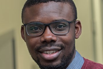 A smiling man wearing glasses and a burgundy sweater, with a collared shirt underneath, is sitting in an indoor setting.