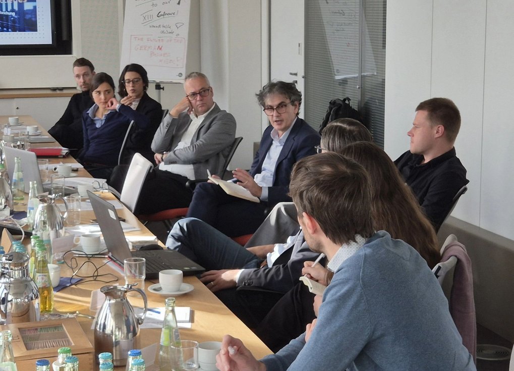 A group of professionals sit around a conference table in discussion. Laptops, notebooks, and drinks are on the table, indicating a collaborative meeting.