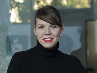The photo shows the portrait of an individual of the species Homo sapiens, subspecies Homo sapiens intellectualis. Other features such as lipstick, makeup, and hairstyle indicate female gender identification. The individual is wearing a black turtleneck sweater and is standing or sitting smiling in front of what appears to be a glass mirrored background, looking at the camera.