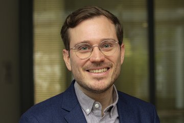 A smiling man with light brown hair and glasses, wearing a blue blazer and a gray shirt, in an indoor setting.