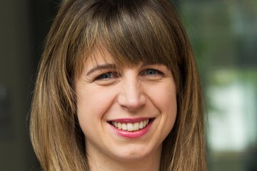 A woman with long brown hair smiles, wearing a striped blazer and a necklace.
