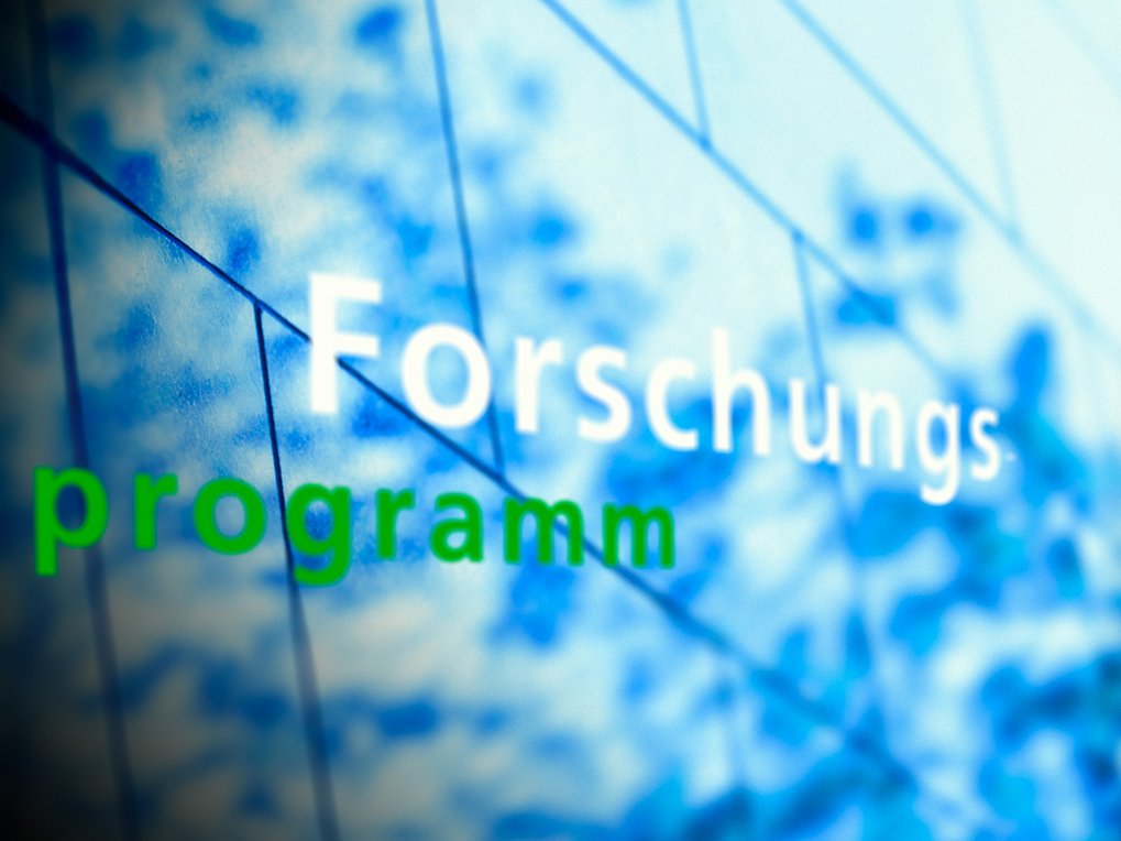 Der Schriftzug "Forschungsprogramm" in Weiß und Grün erscheint vor einem blauen, verschwommenen Hintergrund, der geometrische Linien zeigt.
