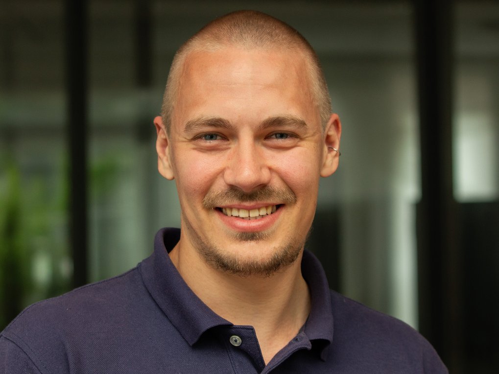 Eine Person in einem dunkelblauen Polohemd steht vor einem Gebäude mit großen Glasfenstern, Pflanzen im Hintergrund.