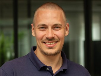 Eine Person in einem dunkelblauen Polohemd steht vor einem Gebäude mit großen Glasfenstern, Pflanzen im Hintergrund.